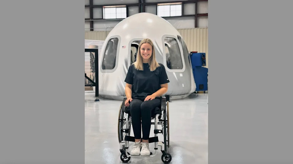 a young woman in a wheelchair sits in front of a white space capsule inside a large hangar