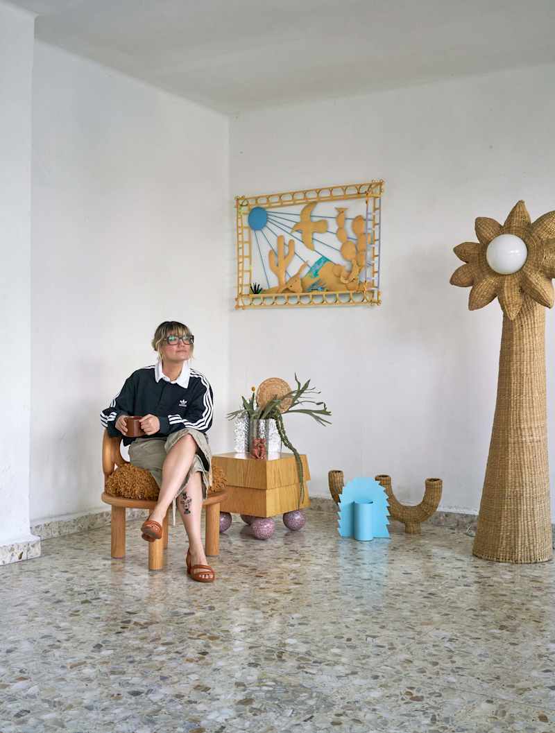 A person sitting in a wooden chair, holding a mug, with a decorative wall art, plants, and unique decor items around.