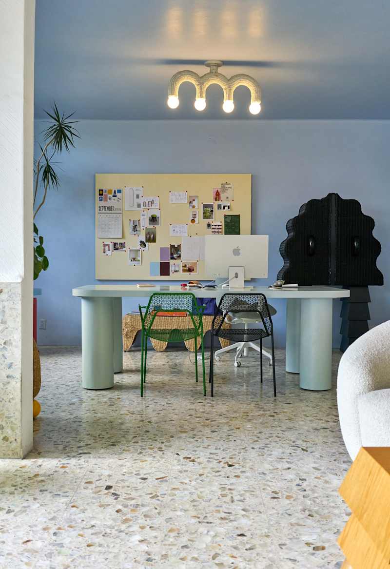 Modern home office with a light blue wall, a large desk, colorful chairs, a bulletin board, and a unique light fixture.