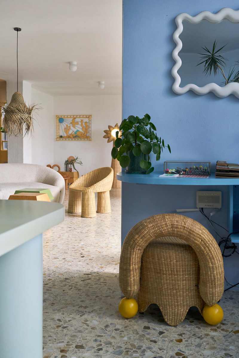Cozy living space featuring a light blue wall, woven furniture, a potted plant, and decorative art.